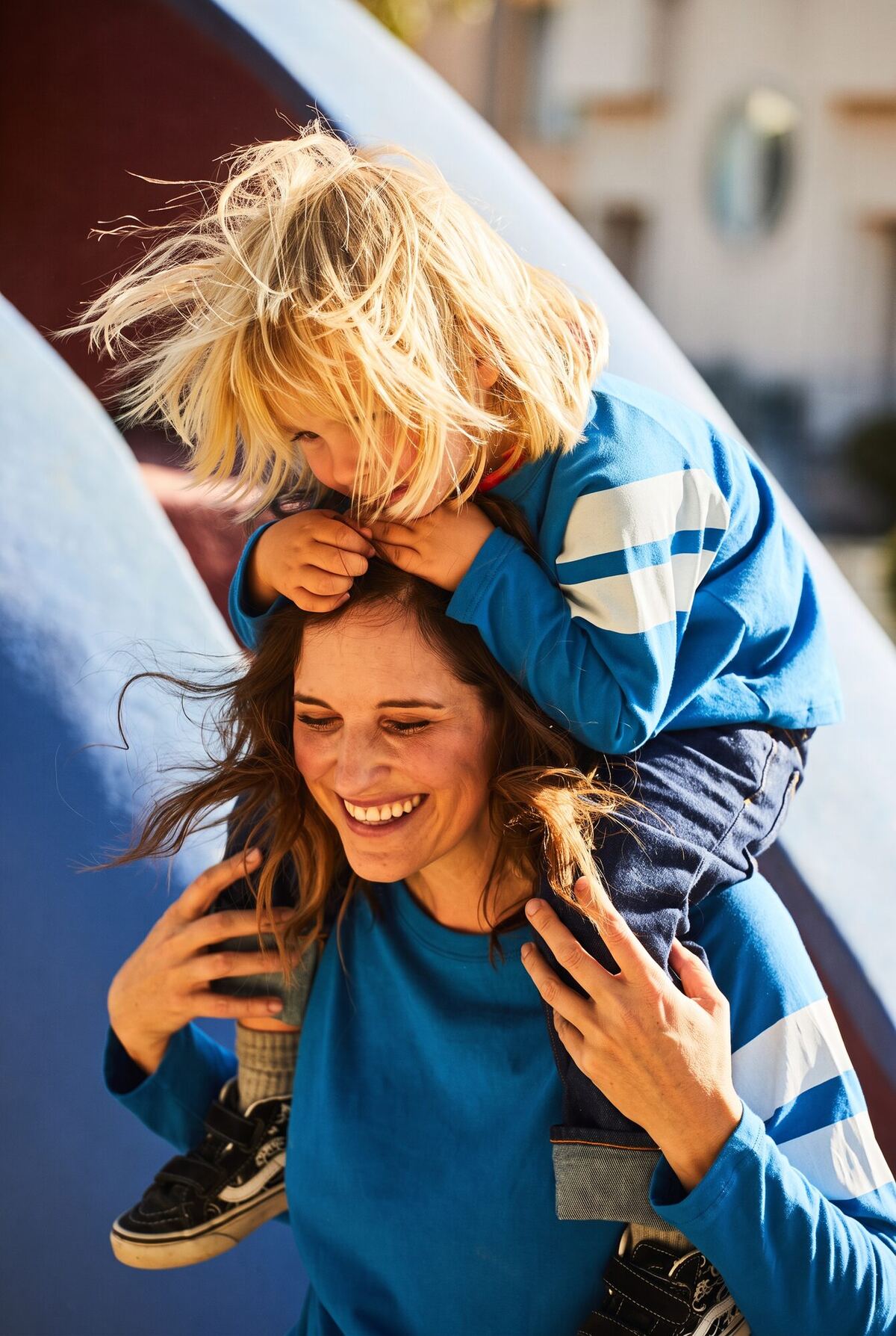 Mallyce laughing with her son on her shoulders at a playground, his blond hair falling over his face